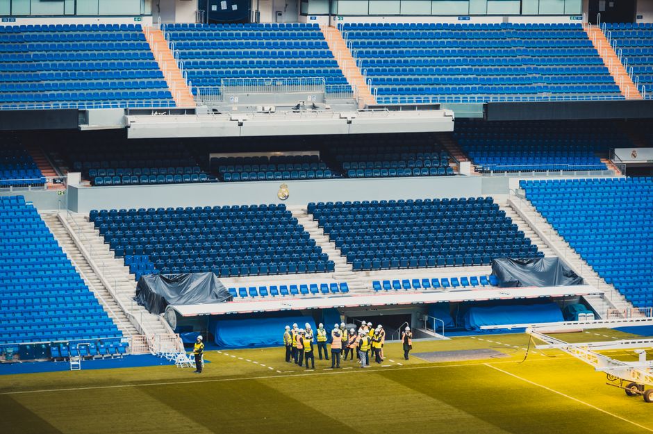 A group of maintenance workers wearing safety helmets and high-visibility vests standing on the grass pitch of Twickenham Stadium, which features tiered seating with blue and orange seats, some sectio