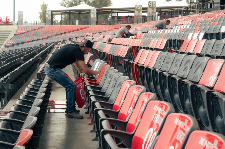 A group of maintenance workers wearing safety helmets and high-visibility vests standing on the grass pitch of Twickenham Stadium, which features tiered seating with blue and orange seats, some sectio